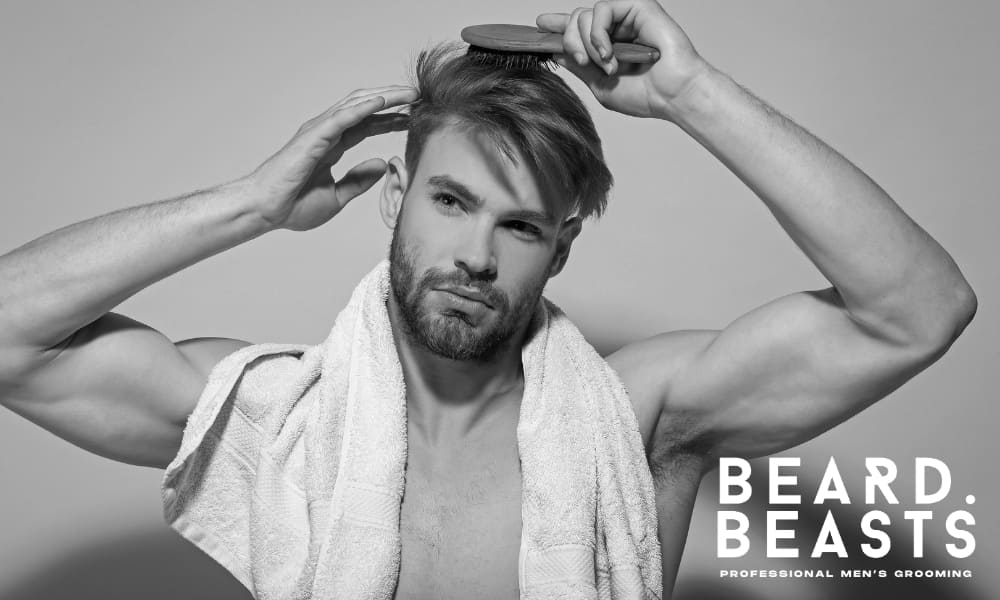 A black-and-white image of a muscular man brushing his hairstyle to one side. He has a well-groomed beard and a confident expression, focusing on styling his hair. A towel is draped over his shoulders, suggesting a post-shower grooming routine. His toned arms and defined features add to the strong, masculine aesthetic.