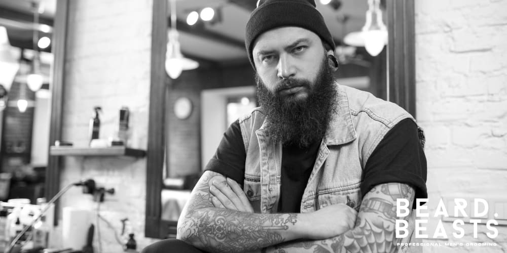 Black and white image of a bearded man with tattoos, sitting confidently in a barber shop. He wears a black beanie, a light denim vest over a black t-shirt, and displays a rugged yet professional demeanor. The vintage-inspired barber shop setting features exposed brick walls, vintage light fixtures, and classic grooming tools in the background.