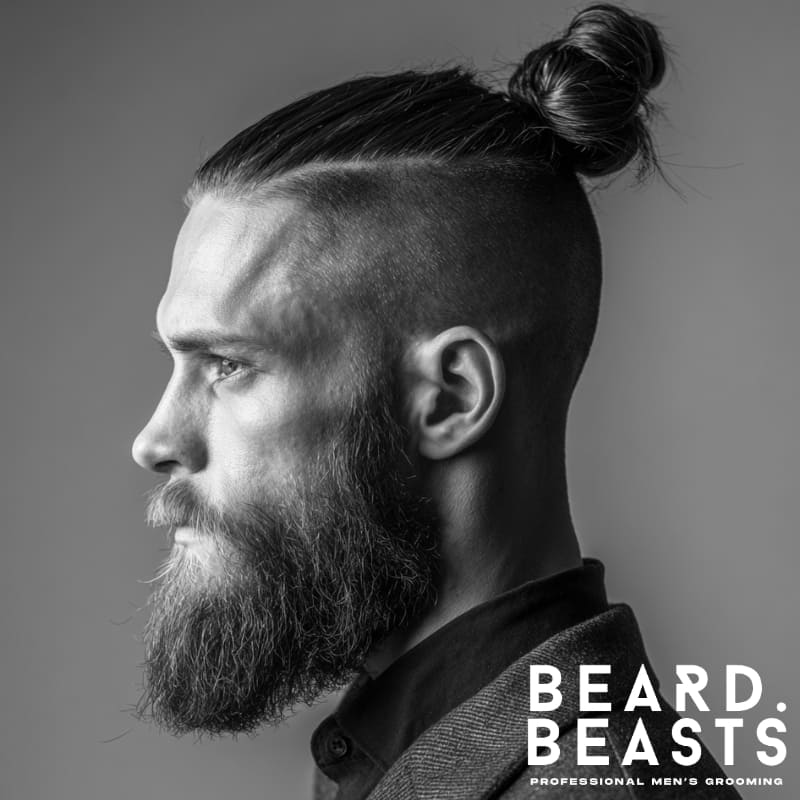Man with straight hair styled into a mid-to-high man bun, high skin fade on the sides, and a full, well-groomed beard in a side profile studio portrait.