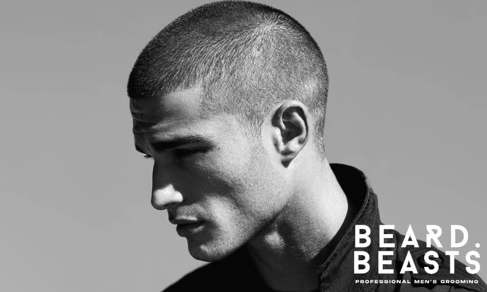 Profile shot of a man with a clean burr cut, showcasing a sharp, uniform hairstyle. His intense gaze and defined features are highlighted by the minimalistic style, complemented by a black jacket for a bold, masculine look.