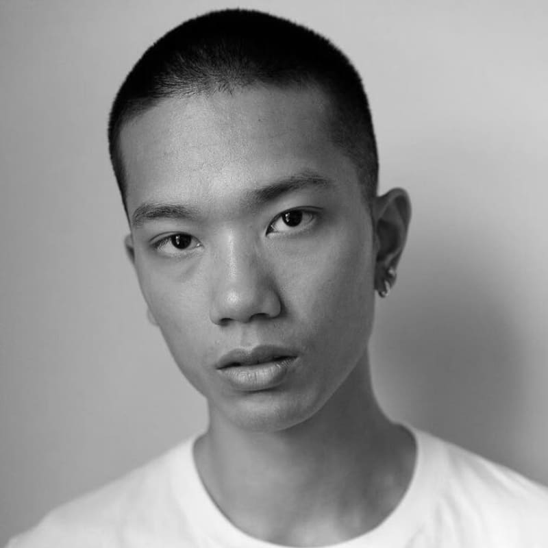 A black and white portrait of a young man with a short, evenly trimmed hairstyle, creating a clean yet textured look. His sharp facial features, including high cheekbones and a defined jawline, are highlighted by the soft, natural lighting. His calm, direct gaze and subtle expression add depth and character to the image. The minimalist background and simple white t-shirt create a timeless, understated aesthetic. A small earring adds a touch of individuality to his effortlessly stylish appearance.