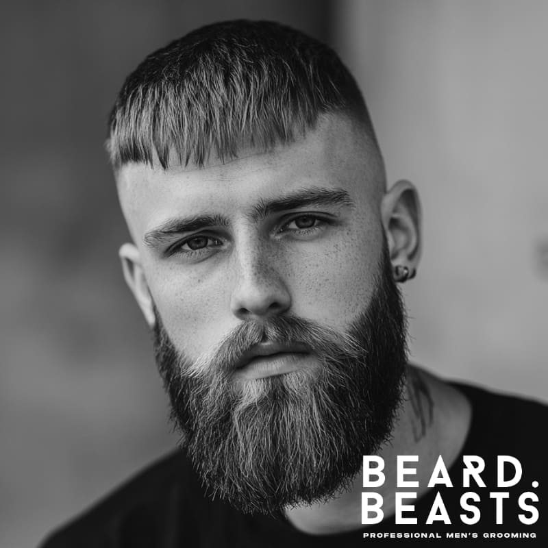 Close-up of a man with a classic high fade crop top hairstyle and full beard, featuring a straight fringe and sharp faded sides.