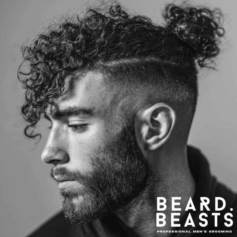 Man with defined curly hair styled into a loose man bun with a high fade and prominent front curls, paired with a full beard in a studio setting.