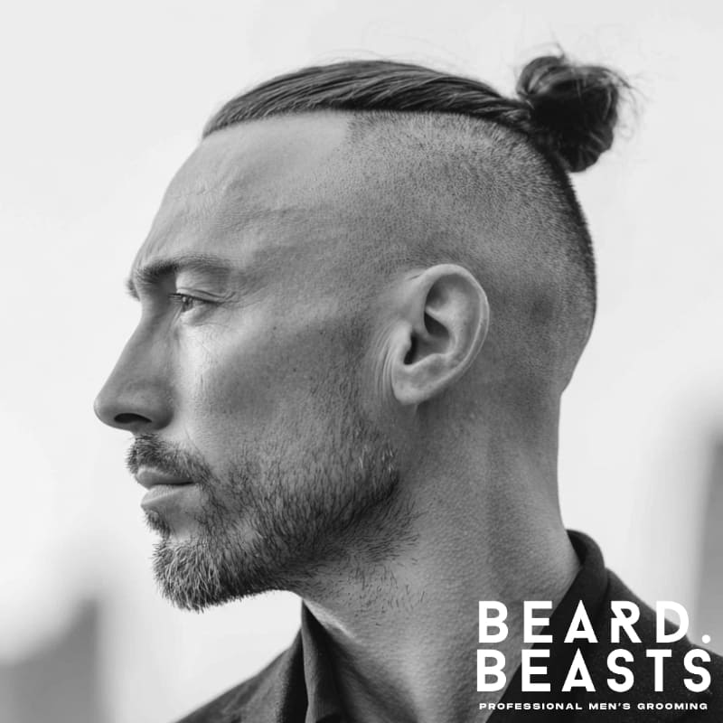 Man with straight hair styled into a slick high man bun, featuring a disconnected high skin fade with no taper, and light stubble beard in a city backdrop.