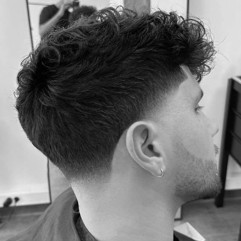 A black-and-white close-up of a man with a low taper fade haircut, featuring a smooth and precise fade that starts just above the ears and blends seamlessly into the neckline. The top is styled with natural, voluminous curls, creating a modern contrast between texture and the clean fade. His sharp beard lineup complements the haircut, enhancing the overall polished look. The barbershop setting in the background reinforces the professional styling and high-quality grooming.