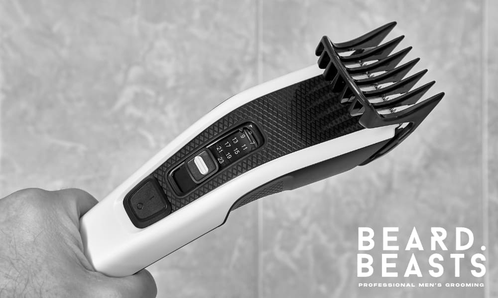 A close-up black and white photograph of a high-quality electric hair clipper, held firmly in a hand. The clipper features an adjustable length setting dial and a sturdy comb attachment, designed for precise and even trimming. The textured grip and sleek design highlight its professional-grade construction. The background is softly blurred, focusing attention on the clipper’s details. The Beard Beasts logo in the bottom right corner reinforces the brand’s commitment to professional grooming tools and high-quality styling.
