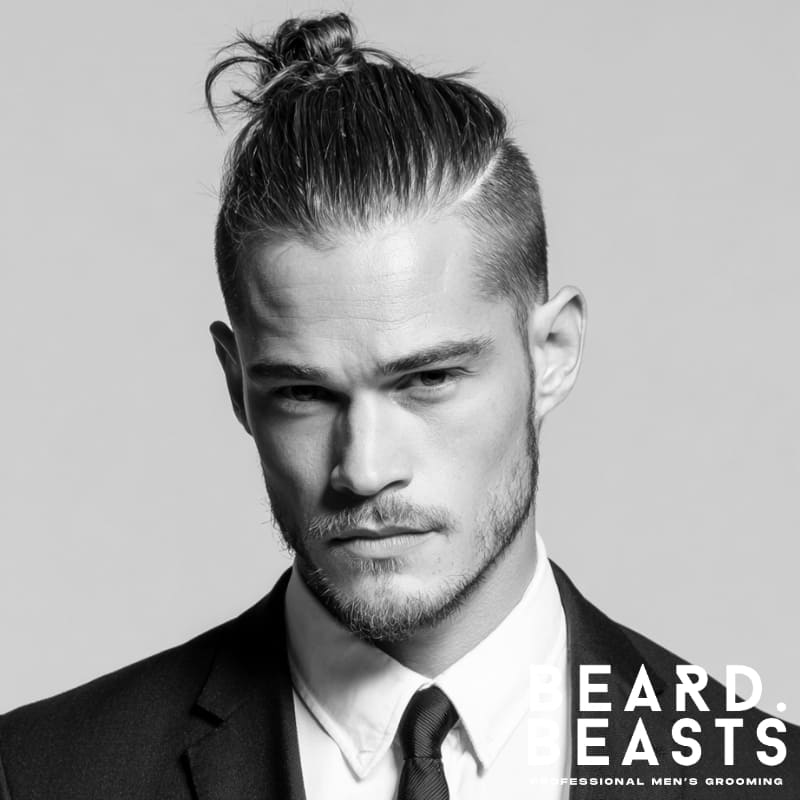 Man with straight hair styled into a slicked-back man bun, featuring a high fade and a sharp hard part line, wearing a suit and light beard.