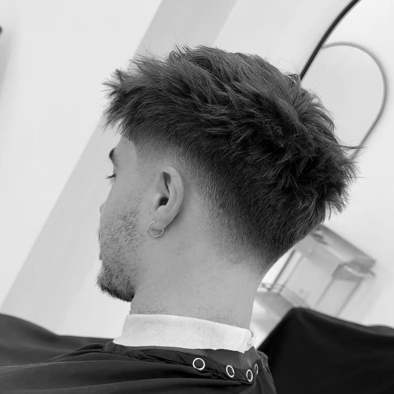 Black and white close-up of a low fade haircut. The hair on the sides and back transitions smoothly from a very short, almost skin fade just above the ear, to longer, messily textured hair on the crown. A subtle stubble beard is visible.