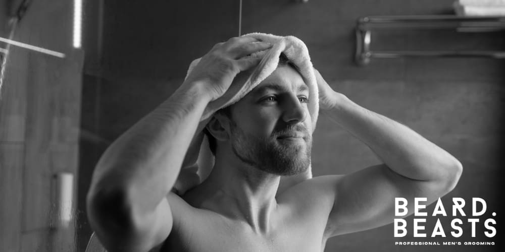 Black and white image of a man drying his hair with a towel in a modern bathroom. The man has a well-groomed beard and appears relaxed, highlighting a grooming routine. The Beard Beasts logo in the corner emphasizes the brand's focus on professional men's grooming and self-care.