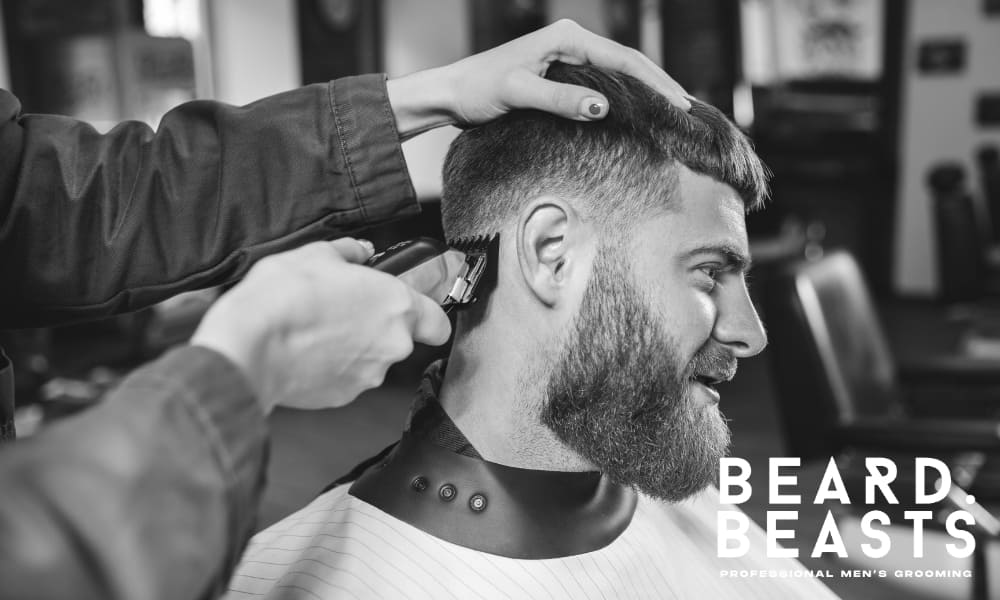 A black-and-white photograph of a bearded man receiving a haircut at a barbershop. The barber is using clippers to create a smooth transition on the sides while holding the client's hair in place for precision. The man appears relaxed and smiling, enjoying the grooming experience. The background features classic barbershop elements, including chairs and mirrors, adding to the professional atmosphere.