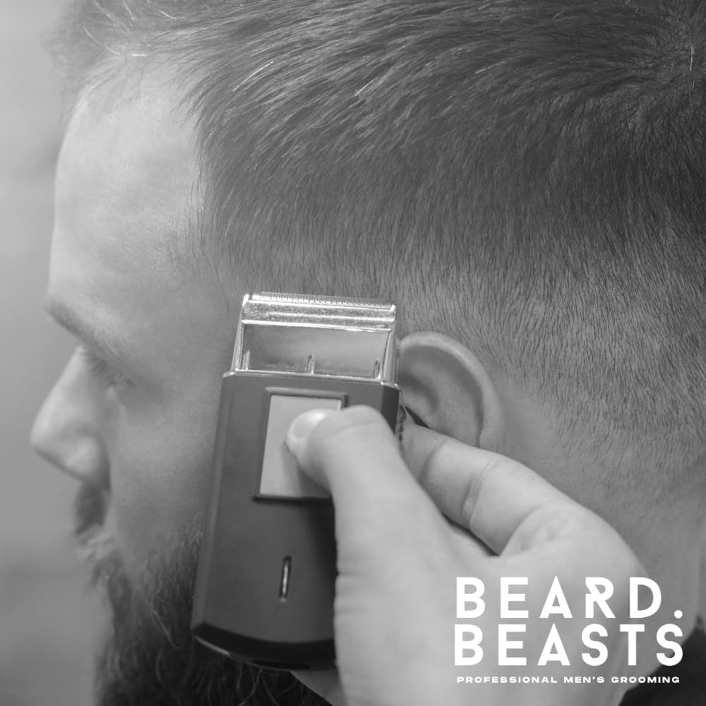 Black and white image of a man receiving a professional haircut at a barbershop. A barber uses an electric foil shaver to create a clean, precise fade around the side of the man's head. The haircut features a smooth transition from longer hair on top to a closely shaved finish near the ear.