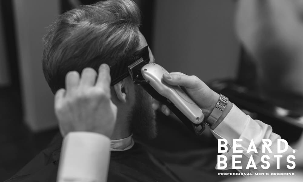 Black and white image of a man getting a cut at a barbershop. A barber uses clippers and a brush to trim the side of his head with precision. The man has a neatly styled look and a well-groomed beard. The Beard Beasts logo appears in the bottom corner, highlighting a professional grooming environment.