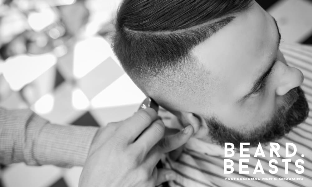 A black-and-white close-up image of a man getting a hard part haircut at a barbershop. The barber is using clippers to refine the sharp fade on the sides, while the top remains neatly combed to one side. The hard part is precisely shaved, adding definition and structure to the hairstyle. The man has a well-groomed beard, enhancing his polished and masculine look.