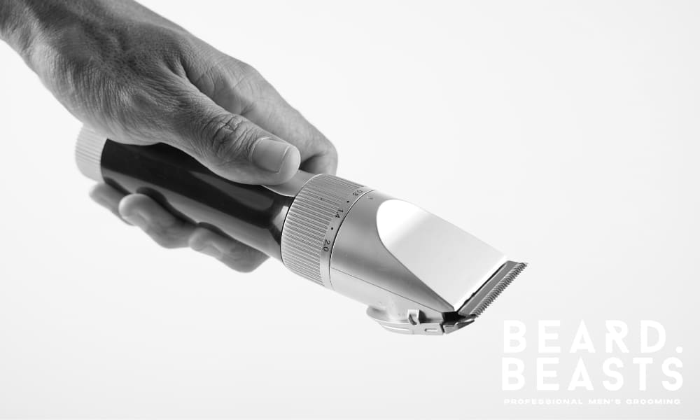 A black and white close-up photograph of a high-quality hair clipper being held by a man's hand. The sleek clipper features an adjustable length dial and sharp precision blades, ideal for creating clean fades and professional haircuts. The minimalistic white background enhances the modern and premium design of the grooming tool. The image highlights the importance of using professional-grade equipment for maintaining sharp hairstyles.