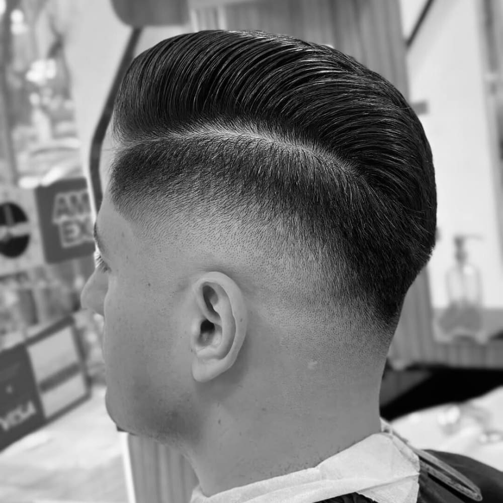 "Black and white image of a man with a sharp side part pompadour haircut in a barbershop setting. The hairstyle features a clean, defined side part with the top styled into a sleek and voluminous pompadour. The sides are tapered into a smooth fade, showcasing a seamless blend from the skin to the longer hair on top. The overall look is polished and professional, emphasizing the contrast between the structured top and the clean, faded sides.