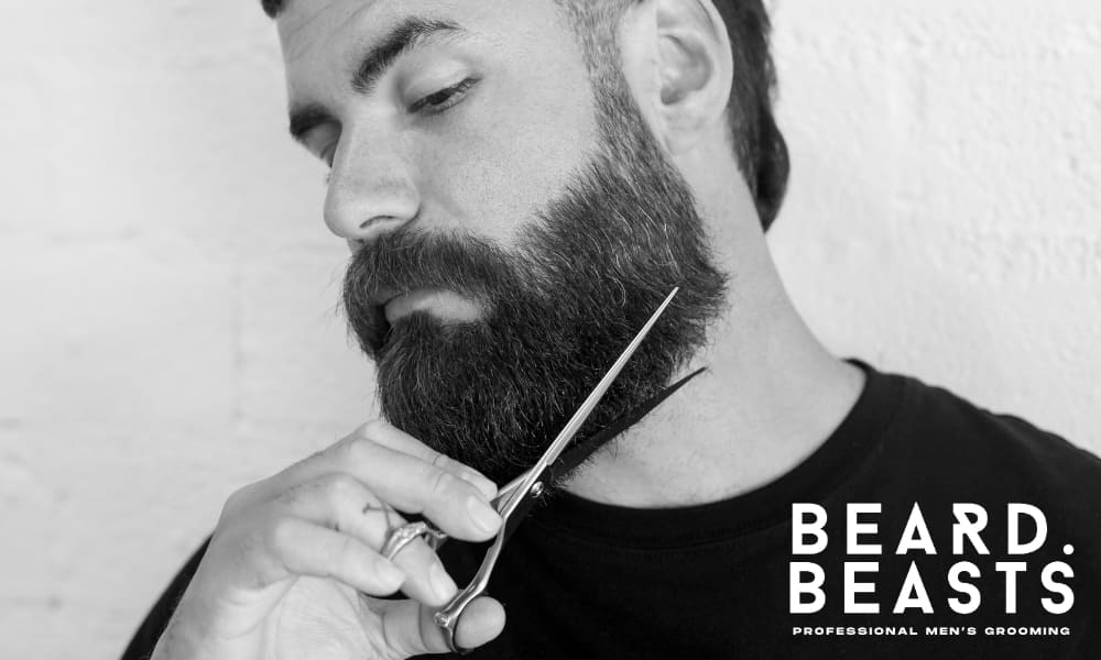 Black-and-white image of a man carefully trimming his thick, full facial hair with a pair of grooming scissors. He holds the scissors at an angle to shape the lower edge of his facial hair, with a focused and precise expression. The man has a neatly styled haircut and is wearing a black t-shirt.