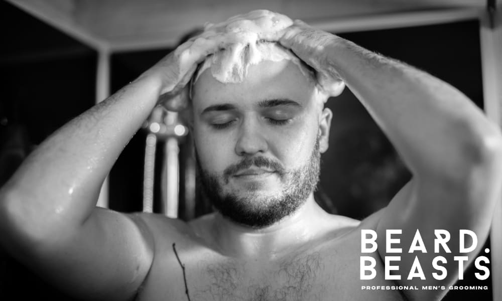Black-and-white image of a man washing his hair in the shower. He has his eyes closed and both hands massaging shampoo into his scalp, with thick lather covering his head. Water droplets are visible on his shoulders, and he has a short, well-groomed beard, conveying a clean and refreshing grooming routine.