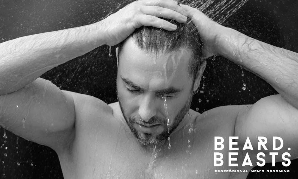 Black and white image of a man washing his hair under running water, with both hands massaging his scalp. Water flows down his face and shoulders, capturing a moment of personal grooming. The Beard Beasts logo appears in the corner, emphasizing professional men’s grooming and hair care.