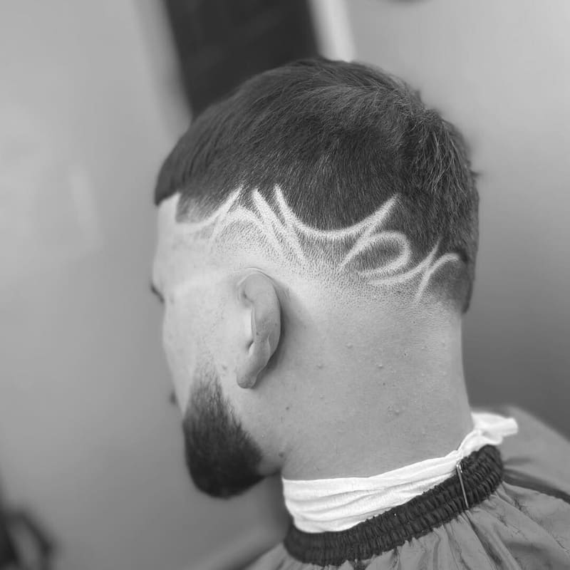 A black-and-white image of a man with a high drop fade with a design. The haircut features a clean, high fade that smoothly tapers around the sides and back, blending into the skin. The lower fade section is enhanced with an intricate shaved design, featuring sharp, curved patterns that add a bold, artistic touch. The top is kept short and neatly trimmed, complementing the structured look of the fade. The background shows a barbershop setting, highlighting the professional craftsmanship of the haircut.