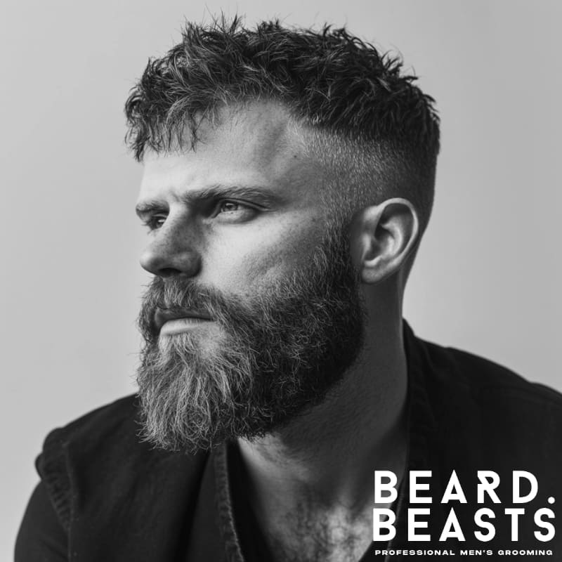 black and white portrait of a bearded man with a short textured crop and a high fade haircut. The top is styled with natural texture, giving it a slightly tousled look, while the high fade creates a sharp contrast on the sides. His full, well-groomed beard complements the rugged yet stylish appearance. He wears a black shirt, adding to the masculine and modern aesthetic.