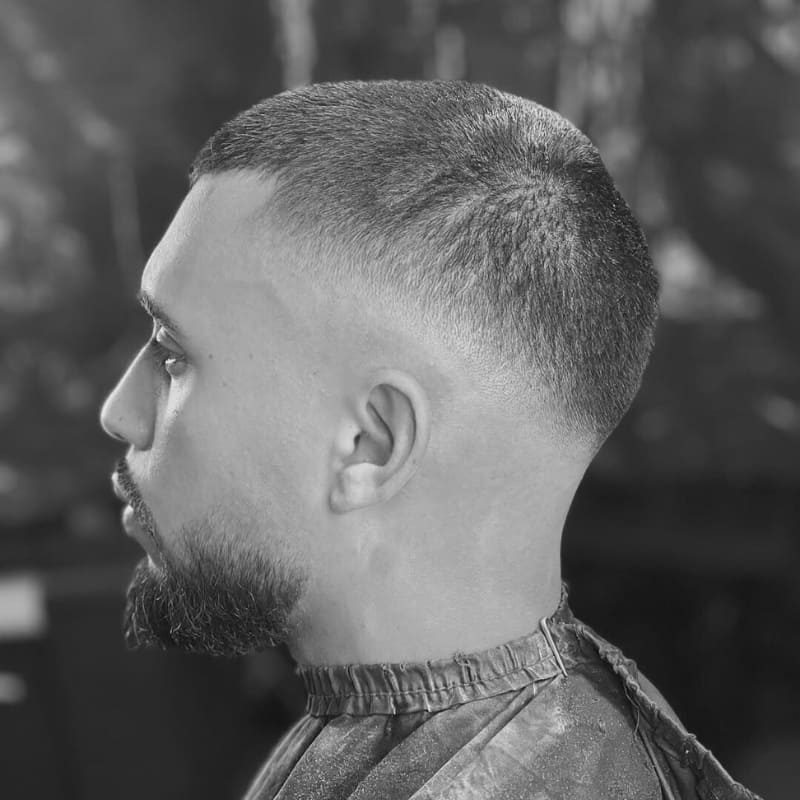 A man with a fresh buzz cut featuring a mid drop fade, seamlessly blending from closely shaved sides into a slightly longer, even-length top. The crisp fade curves naturally around the head, complementing his well-groomed beard for a sharp and polished look. The black-and-white filter enhances the contrast, highlighting the precision of the cut. The soft lighting and blurred background create a professional and stylish barber shop aesthetic.