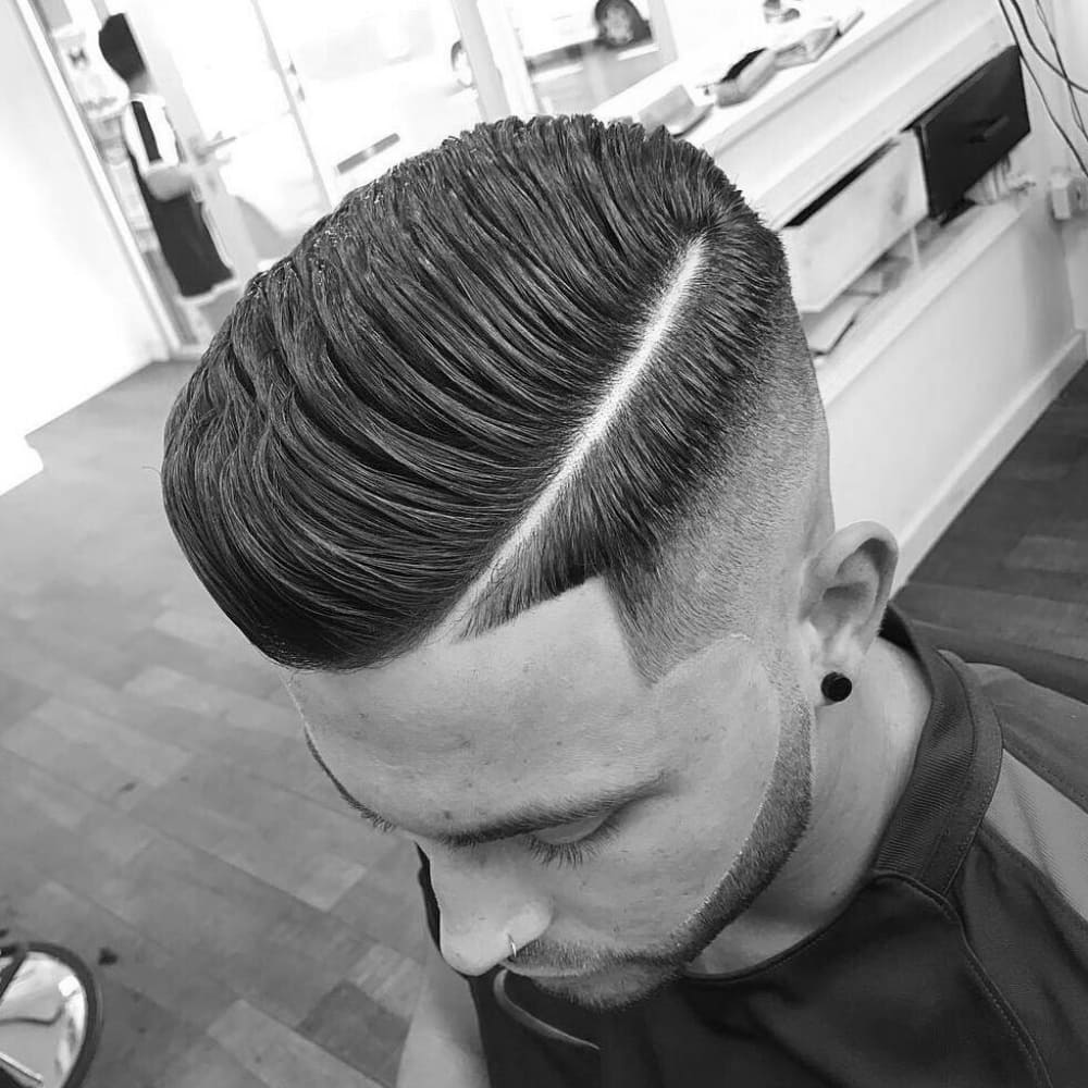 Black and white image of a man with a sharply defined side part pompadour hairstyle. The haircut features a precise, clean side part with the top styled into a voluminous, combed-back pompadour. The sides are expertly faded, creating a strong contrast with the longer hair on top. The man's grooming is immaculate, with a well-shaped beard that complements the hairstyle. The barbershop setting and natural lighting emphasize the crisp lines and polished look of this classic yet modern men's hairstyle.