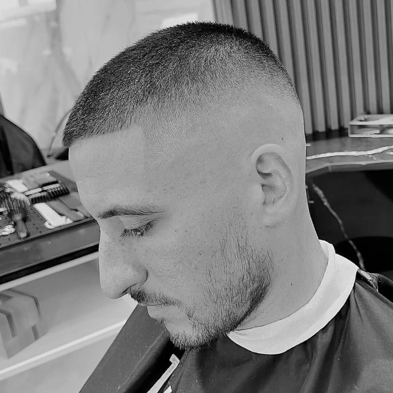 A man with a fresh buzz cut featuring a high drop fade, showcasing a clean and precise blend. The haircut transitions seamlessly from a closely shaved fade into slightly longer textured hair on top. His sharp line-up enhances the crisp and structured look. The monochrome filter highlights the contrast between the fade and the textured top. In the background, barber tools are neatly arranged, reinforcing the professional grooming setting.