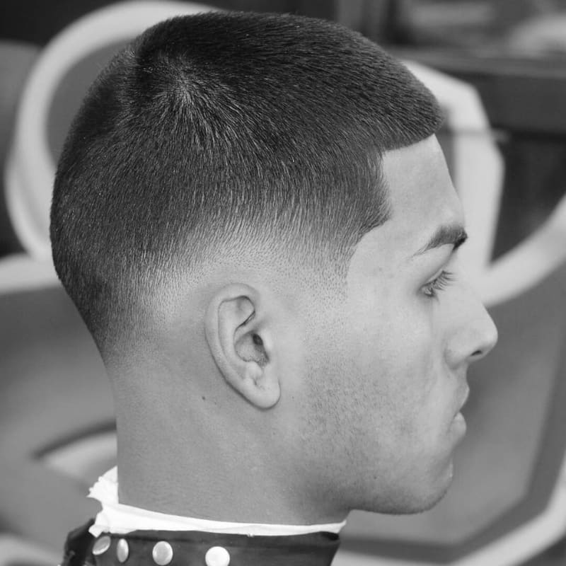 A man with a fresh buzz cut featuring a low drop fade, seamlessly blending from a closely shaved fade into a slightly longer, even-length top. The fade follows the natural curve of the head, creating a smooth and polished transition. A sharp line-up frames the forehead and temples, enhancing the crisp, clean-cut look. The black-and-white filter highlights the precision of the haircut, with a blurred barbershop background adding to the professional grooming aesthetic.