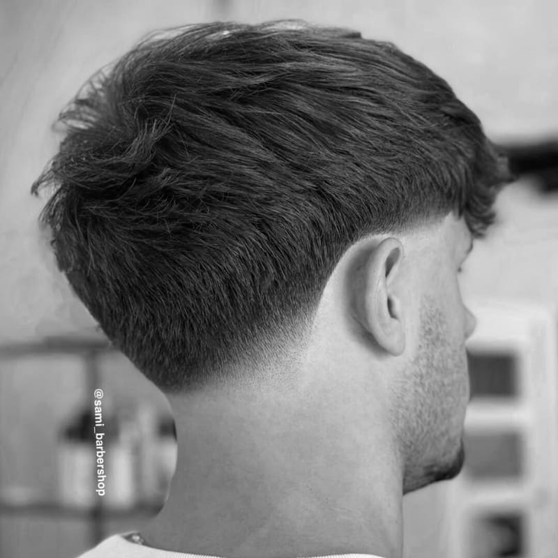 A high-quality black-and-white close-up of a man’s low taper fade haircut. The fade starts just above the ears and gradually blends into the natural hairline at the nape, creating a seamless and polished look. The top features longer, textured hair that adds contrast to the sharp, clean taper. The image captures the precision of the cut, emphasizing the smooth transition and professional finish. The background suggests a modern barbershop setting, reinforcing the focus on high-quality men's grooming.