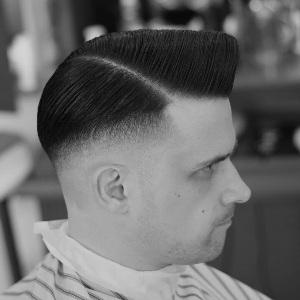 Black and white image of a man with a classic side part pompadour haircut. The hairstyle features a sharp, defined side part with a sleek, voluminous pompadour on top. The sides are tapered with a clean fade, highlighting the contrast between the neatly styled top and the short, well-groomed sides. The man's appearance is professional and polished, sitting in a barbershop setting with a barber cape draped around his shoulders.