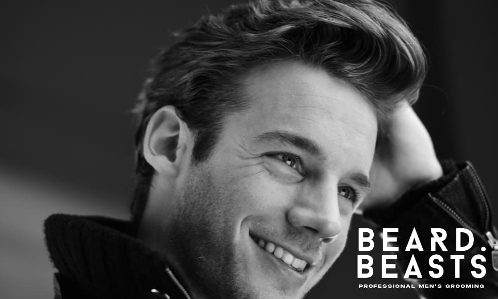 Black and white image of a smiling man with naturally voluminous hair, demonstrating how to add volume to men's hair. His hairstyle features lift and texture, showcasing effective grooming techniques for fuller, thicker-looking hair.