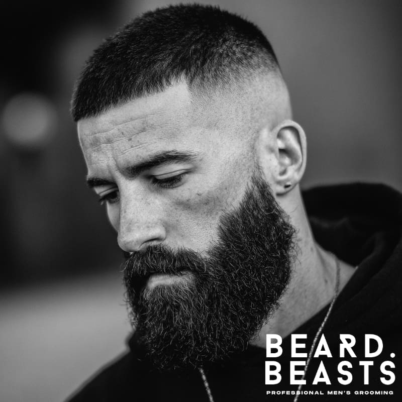 Black and white close-up of a man with a very short buzz cut, a sharp high drop fade, and a thick, well-groomed full beard.
