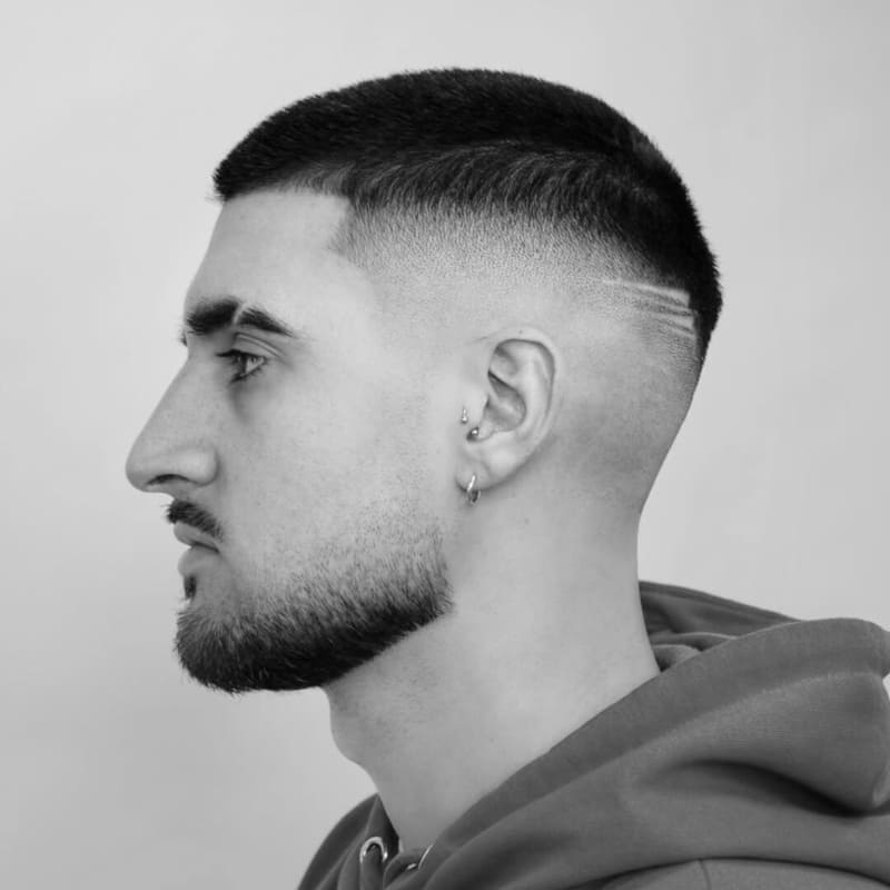 Profile view of a man showcasing a sharp buzz cut paired with a high drop skin fade, featuring a subtle carved line design, and a neatly trimmed beard.
