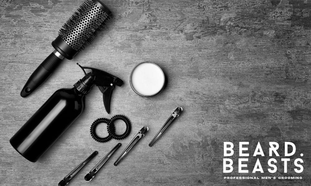 A black-and-white flat lay image featuring men's hair styling tools on a textured surface. The arrangement includes a round brush, a spray bottle, a jar of styling product, hair clips, and coil hair ties, all neatly positioned for a professional grooming setup.