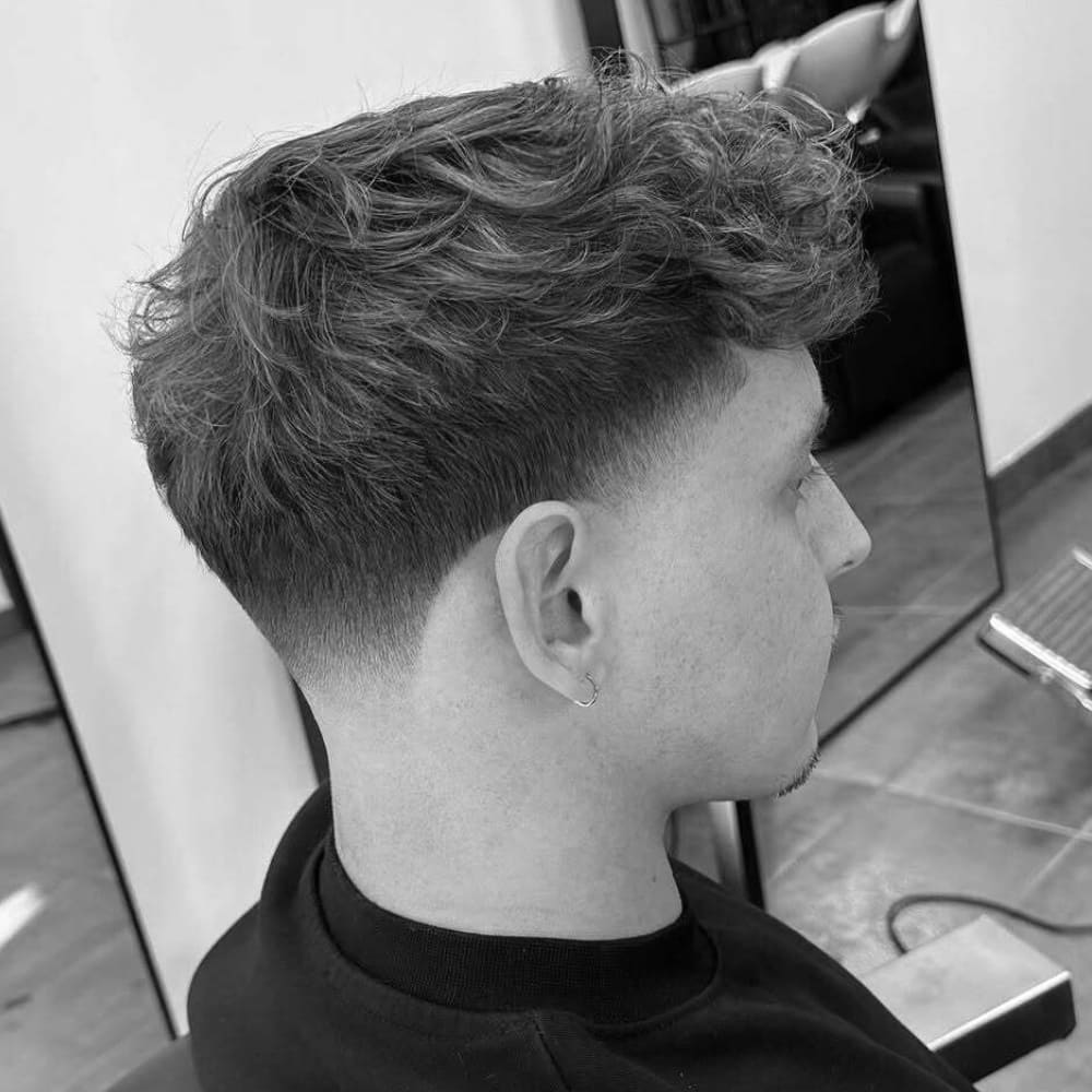 Side profile of a man with a modern messy textured crop hairstyle featuring a low taper fade. The top hair is styled with natural, tousled waves, adding volume and movement. The sides and back blend seamlessly into a clean, gradual fade, highlighting the contrast between the textured top and the sharp, well-groomed sides. The overall look is stylish, contemporary, and versatile, suitable for both casual and professional settings.
