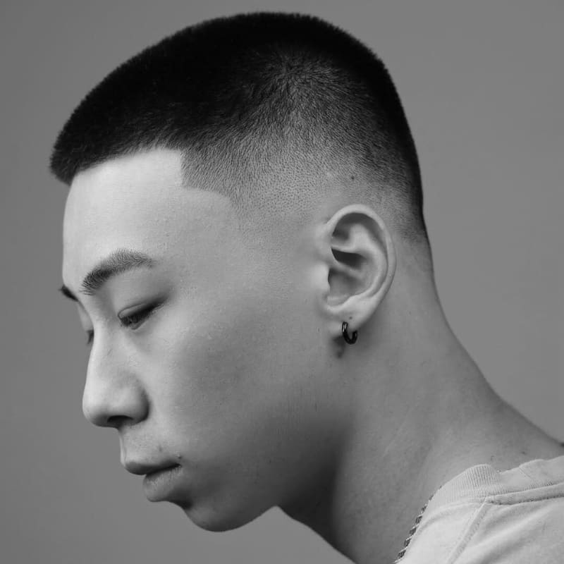 A stylish black and white portrait of an Asian man with a mid fade buzz cut, featuring a sharp, clean hairline and a smooth taper fade on the sides. The precise fade transitions seamlessly into the slightly longer textured top, creating a modern and well-groomed appearance. His subtle accessories, including a small hoop earring and chain necklace, add a touch of personality to the look. The neutral background keeps the focus on the haircut’s sharp details, making it a perfect example of a contemporary, low-maintenance style. This haircut balances professionalism and style, making it ideal for both casual and formal settings.