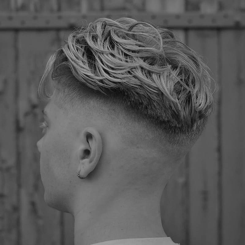A black and white close-up image of a young man showcasing a mid fade undercut with a voluminous, wavy blonde top. The fade starts high on the sides and sharply disconnects from the textured longer hair, creating a bold contrast. The back tapers smoothly towards the nape, while the top is styled in a natural, tousled look. The subject has a small hoop earring and is standing against a rustic wooden backdrop, adding a rugged and stylish feel to the image.