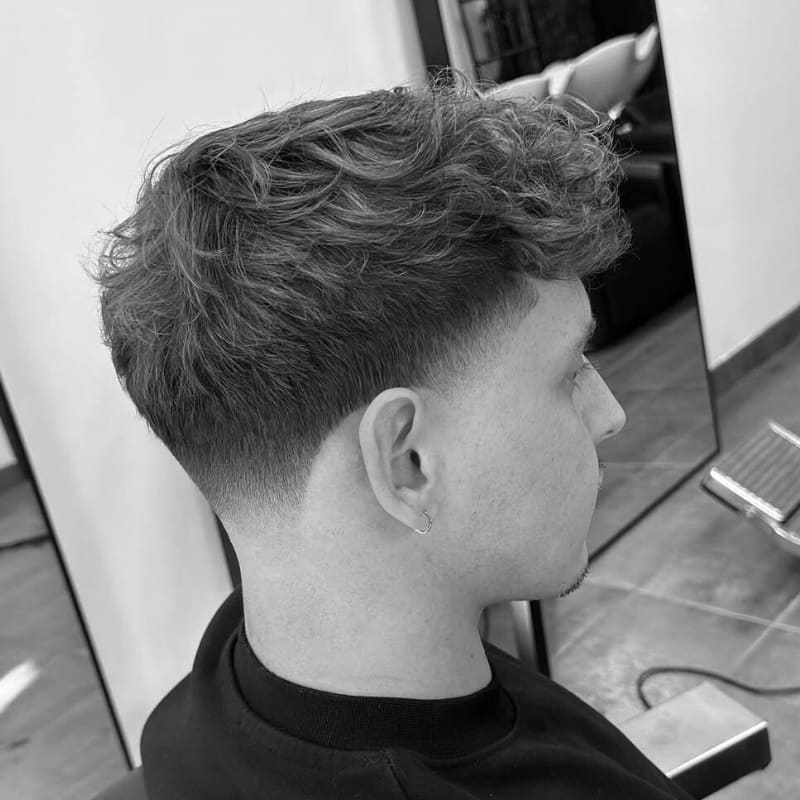 Black and white close-up of a men's taper haircut. The hair on the sides and back gradually shortens from the longer top, maintaining length and not blending to skin at the sideburns and nape. The top features full, curly, and textured styling.