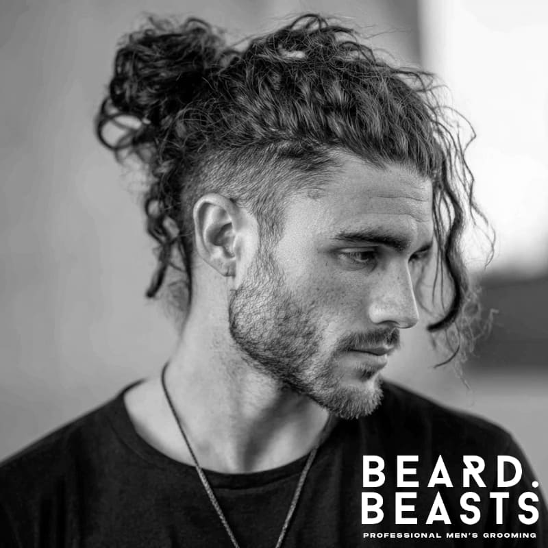 Man with thick, curly hair styled into a loose man bun with a high fade, featuring natural texture and light facial stubble in an outdoor setting.