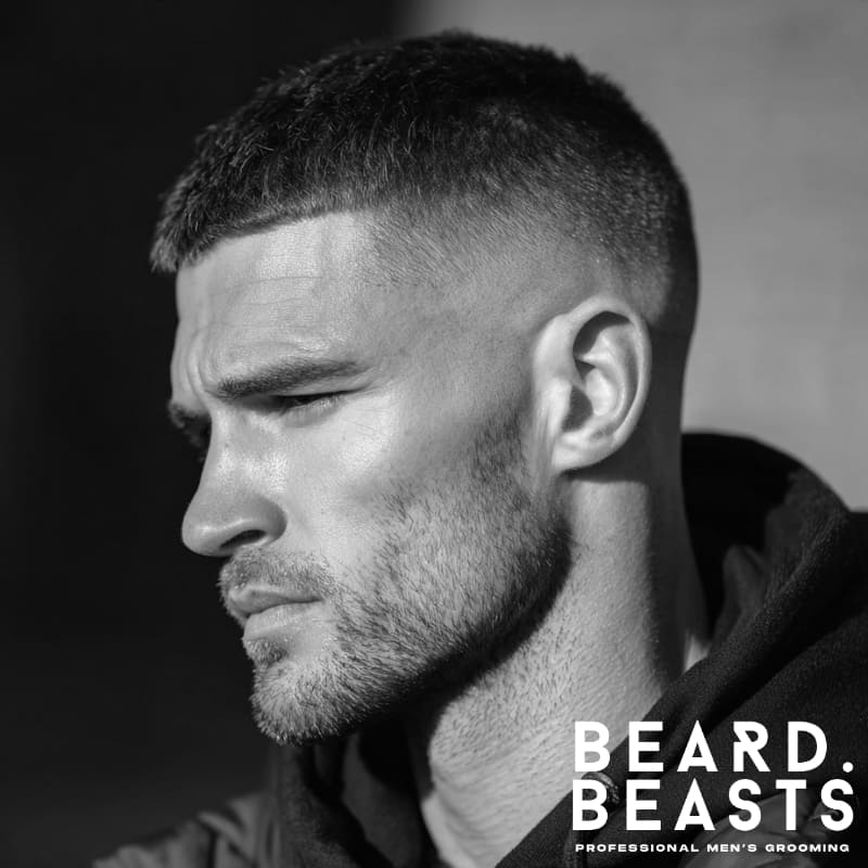 Profile view of a man with a short, textured buzz cut on top, a clean high drop fade on the sides, and light stubble facial hair.