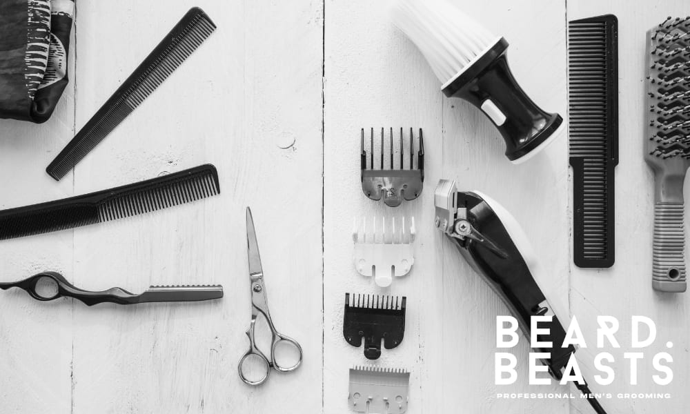 A black-and-white flat lay image displaying men's grooming and hairstyling tools arranged on a white wooden surface. The assortment includes combs, scissors, a hair clipper with multiple guard attachments, a shaving brush, and a textured round brush, creating a professional barbershop aesthetic.