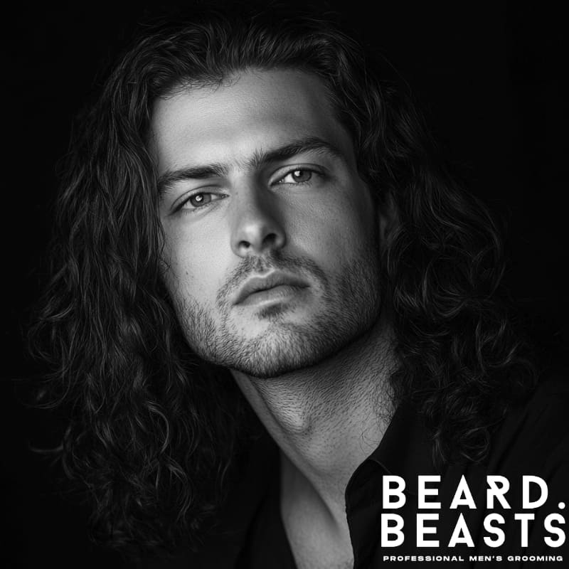 Black and white portrait of a man with long curly hair and medium stubble beard, wearing a black shirt, showcasing the Long Curls and Medium Stubble beard style combo for men – Beard Beasts professional men's grooming.