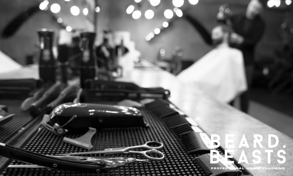 A close-up view of professional grooming tools arranged neatly on a barbershop counter with a blurred barber and client in the background.