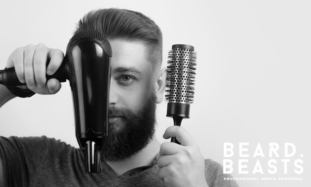Man with a full beard and styled hair holding a hair dryer and round brush in front of his face, promoting men’s grooming tools with Beard Beasts branding.