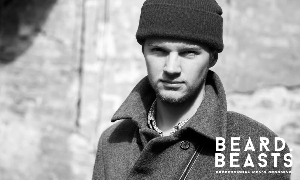 tylish young man with a short beard wearing a wool coat and knit beanie, posing confidently outdoors in black and white – Beard Beasts professional men’s grooming brand image.