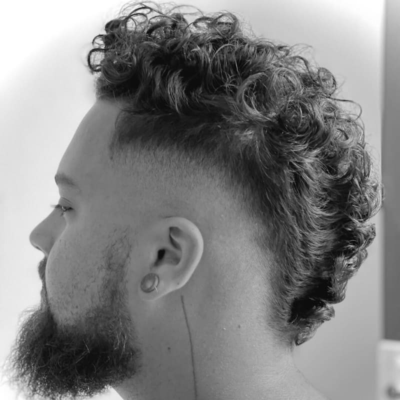 ide profile of a man with tightly curled hair styled in a mohawk, featuring a sharp fade and a full, groomed beard.