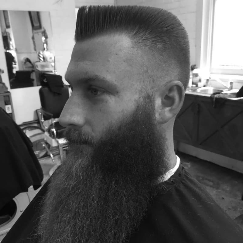 Profile view of a man with a flat top haircut and a long, full beard, featuring sharp lines and a high skin fade in a classic barbershop setting.