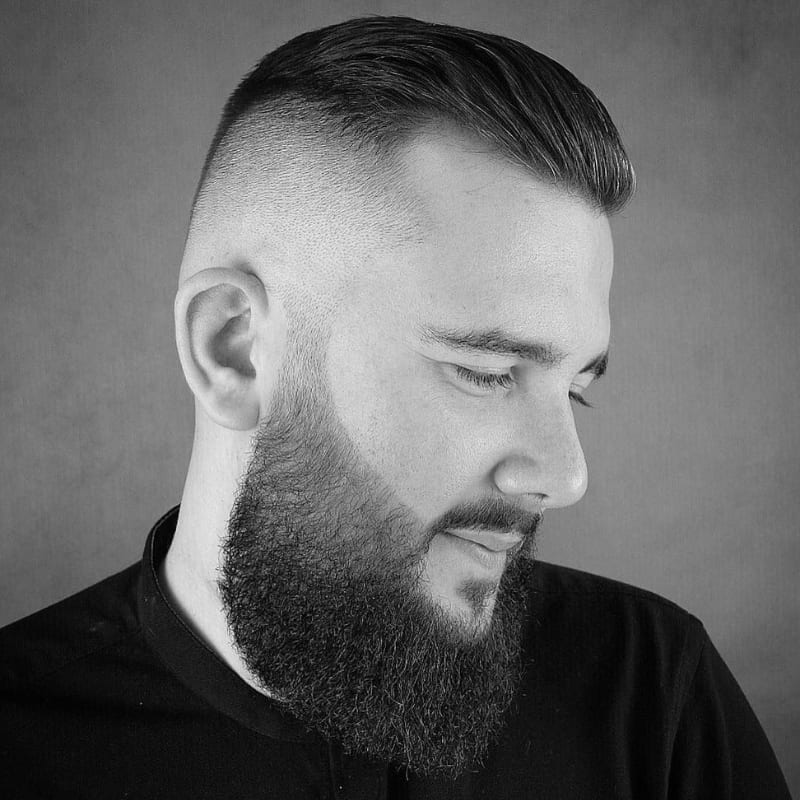 Profile view of a man with a high and tight haircut and a full, well-groomed beard, featuring a sharp skin fade and neatly styled top.