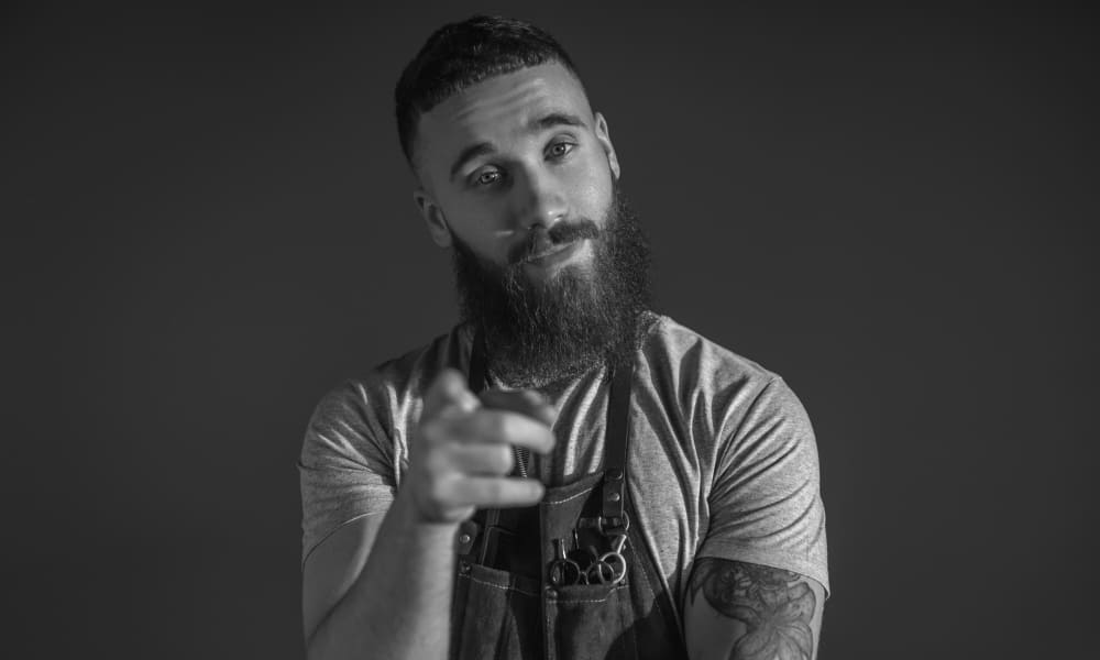 Barber with a full beard and tattoos pointing at the camera, ready to give an Arthur Shelby haircut with a disconnected undercut and slicked-back top.