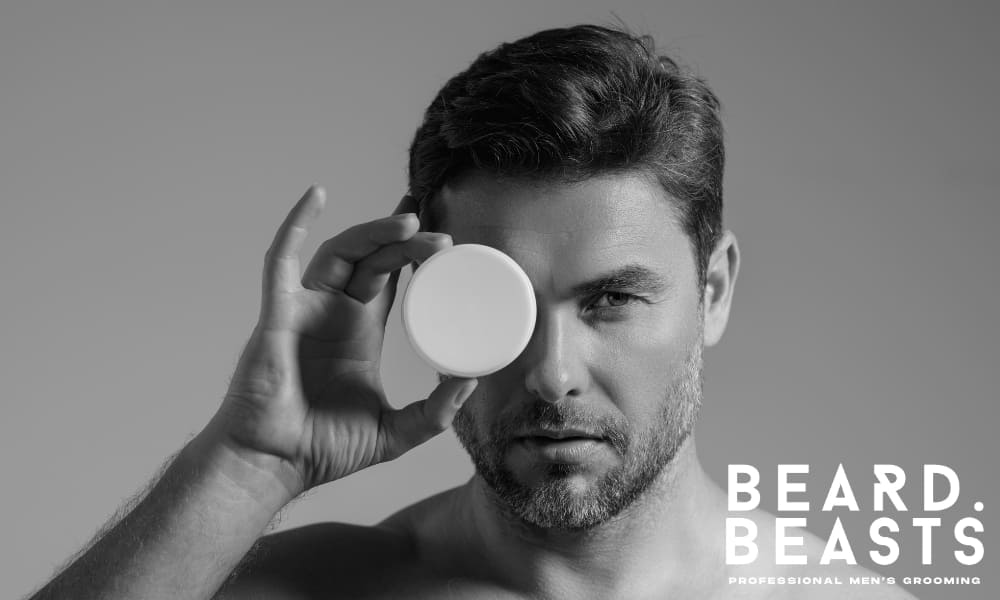 Confident man holding a hair paste container in front of his eye, shot in black and white with soft lighting—symbolizing precise grooming.